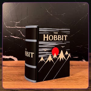 May include: A decorative book-shaped object titled The Hobbit, featuring a black cover with a graphic illustration of beige mountains, a bright red sun, and two white birds against a dark sky. The spine is black with light blue horizontal bands.