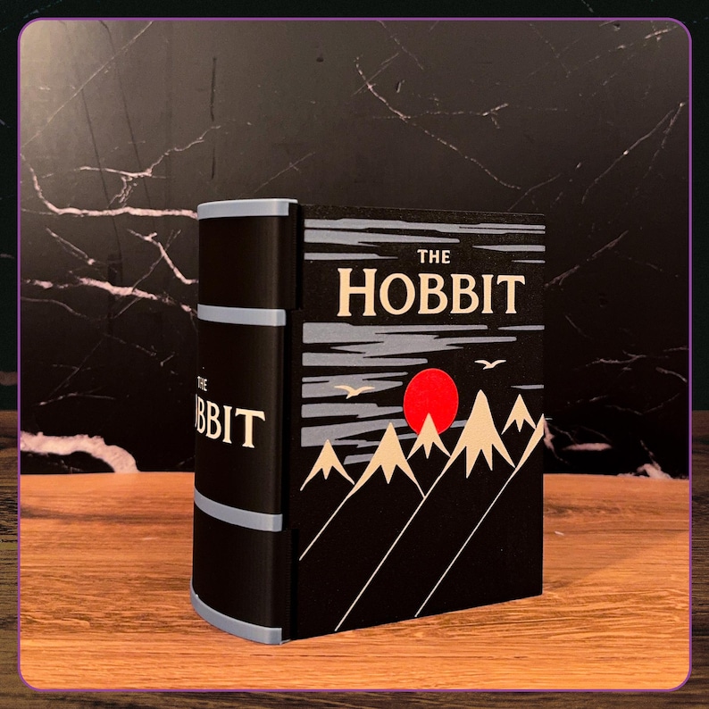 May include: A decorative book-shaped object titled The Hobbit, featuring a black cover with a graphic illustration of beige mountains, a bright red sun, and two white birds against a dark sky. The spine is black with light blue horizontal bands.