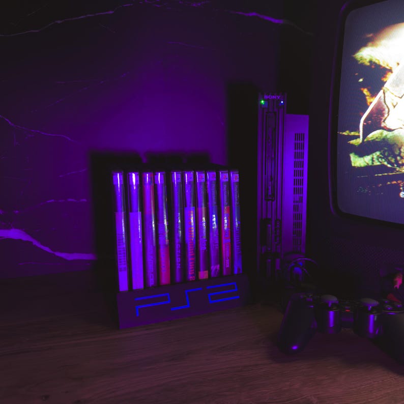 May include: A collection of PS2 game cases in a black holder with a blue neon PS2 logo. A silver PS2 console and a controller are also present. The scene is lit with purple lighting, giving a retro gaming feel.