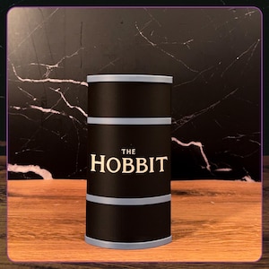 May include: A cylindrical black container with light gray horizontal bands at the top, middle, and bottom. The text THE HOBBIT is printed in a cream-colored serif font on the front center of the black surface of this upright storage canister.