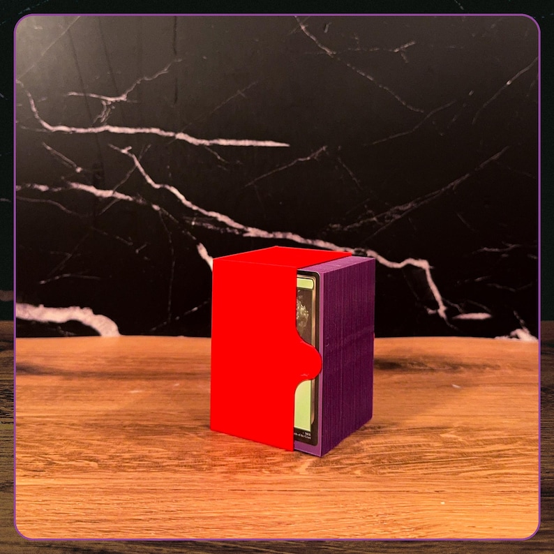 May include: A rectangular deck box featuring a bright red exterior shell and a deep purple inner compartment. The box is designed to hold a stack of trading cards, with a single card visible through the side opening of the purple container.