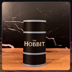 May include: A cylindrical black container with light gray horizontal bands at the top, middle, and bottom. The text THE HOBBIT is printed in a cream-colored serif font on the front center of the black surface of this upright storage canister.