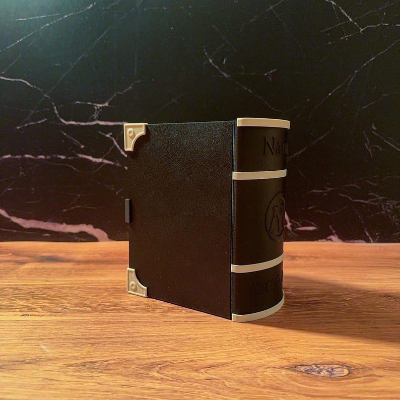May include: A black, book-shaped storage box with a curved spine featuring white horizontal bands and a circular emblem. The cover has light-colored corner protectors and a small latch on the side, presenting a structured, rectangular design.