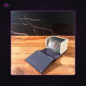 May include: A rectangular deck box with a white exterior and a dark blue, textured folding lid. The box is open, revealing a stack of trading cards with dark, illustrated artwork visible on the front card. The structure is held together by small metal rivets.
