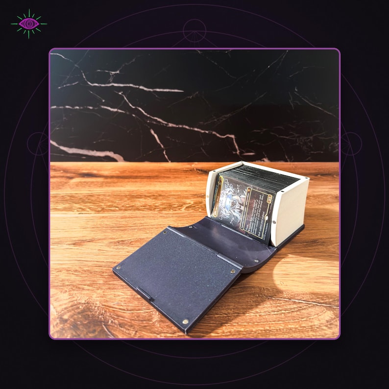 May include: A rectangular deck box with a white exterior and a dark blue, textured folding lid. The box is open, revealing a stack of trading cards with dark, illustrated artwork visible on the front card. The structure is held together by small metal rivets.