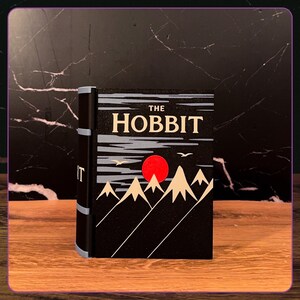 May include: A black book titled The Hobbit with a graphic illustration on the cover featuring a bright red sun, beige mountain peaks, and two white birds against a dark sky. The spine is black with light blue accents and the word Hobbit printed.