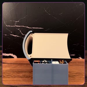 May include: A book-shaped storage container with a black frame and cream-colored pages. A light blue drawer is pulled out from the bottom, revealing several multi-sided gaming dice inside the two compartments of the storage tray.