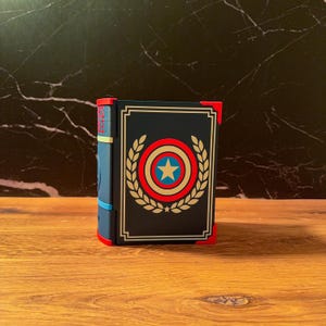 May include: A rectangular storage box designed to resemble a book with a dark blue cover, featuring a central circular emblem with a white star, red and blue rings, and gold laurel leaves. The box has red corner accents and a blue spine.