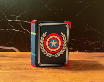 May include: A rectangular storage box designed to resemble a book with a dark blue cover, featuring a central circular emblem with a white star, red and blue rings, and gold laurel leaves. The box has red corner accents and a blue spine.