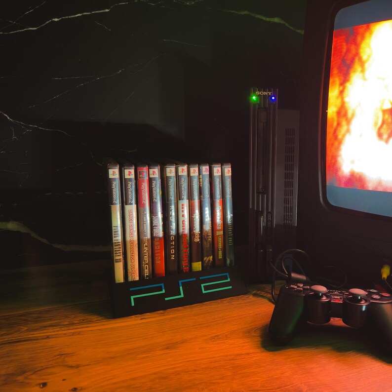 May include: A black PS2 game holder with a neon green and blue design, holding several game cases. A black PS2 console and a controller are also visible, along with a retro television.