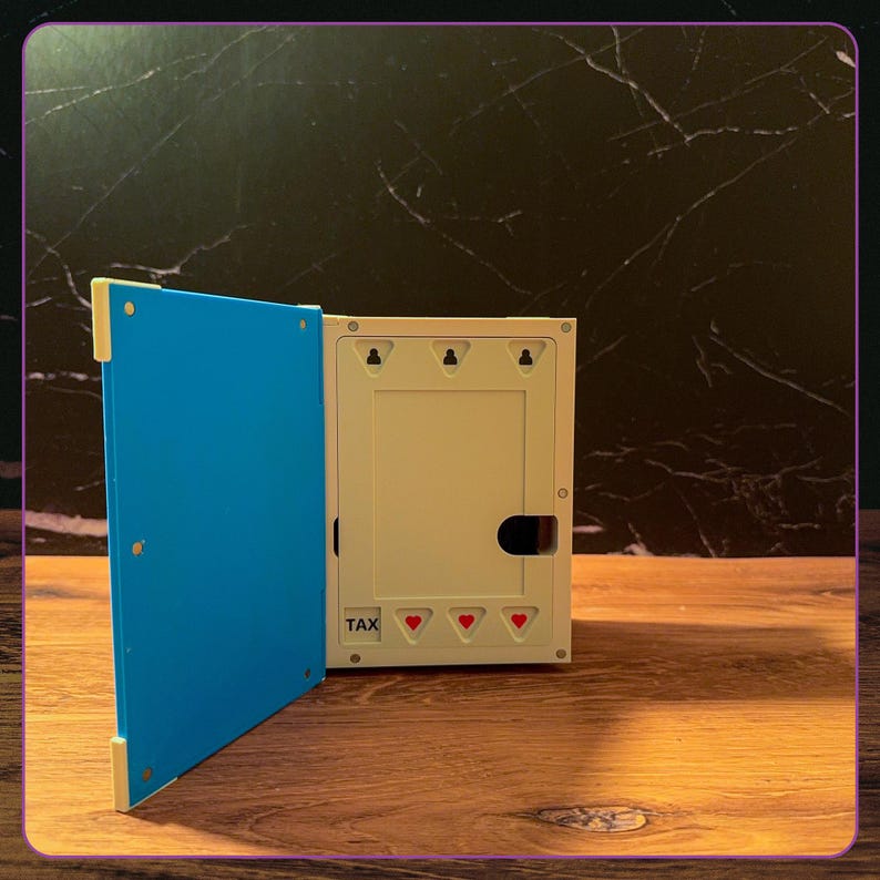 May include: A rectangular white plastic wall-mounted box with a hinged bright blue door. The interior features a central rectangular cutout, three top keyhole slots for mounting, and the word TAX printed above three small red heart icons.