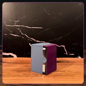 May include: A rectangular deck box for trading cards featuring a light blue outer sleeve with a circular cutout and a deep purple inner container. The box is partially open, revealing a stack of playing cards with white borders inside the structure.
