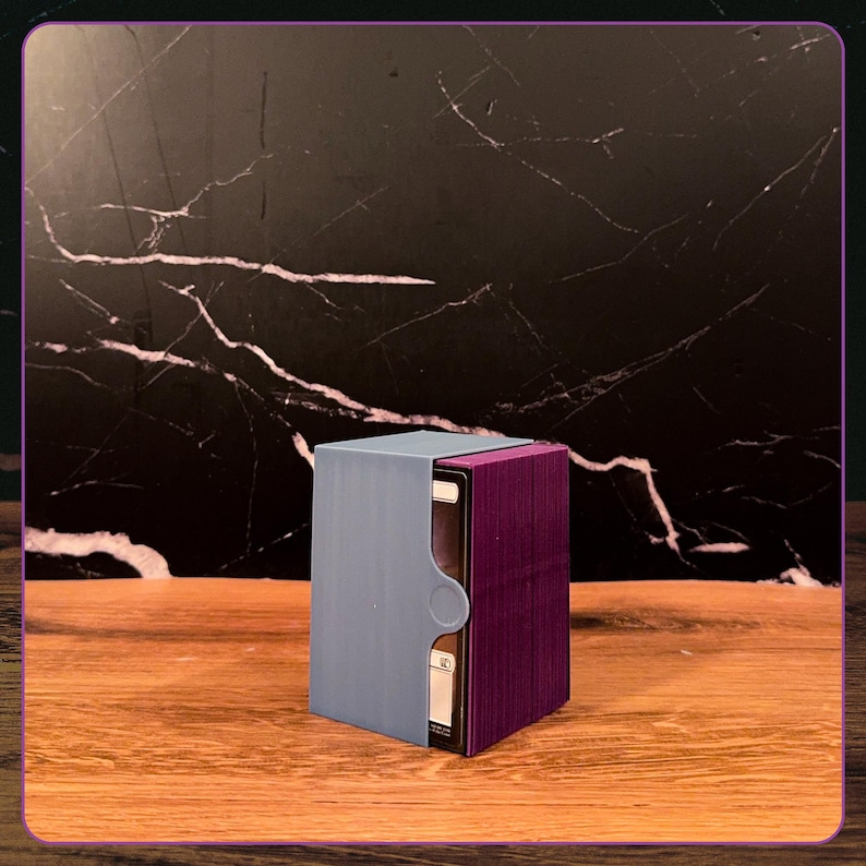 May include: A rectangular deck box for trading cards featuring a light blue outer sleeve with a circular cutout and a deep purple inner container. The box is partially open, revealing a stack of playing cards with white borders inside the structure.