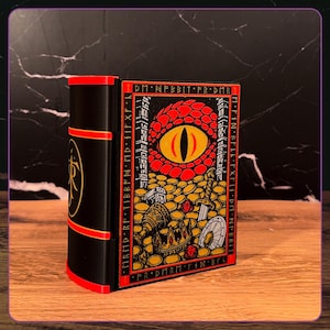 May include: A black rectangular box shaped like a book with red accents. The front panel features a large red dragon eye surrounded by gold coins, a crown, a sword, and a shield. The border contains text reading DM N FBBIT FR DMR.