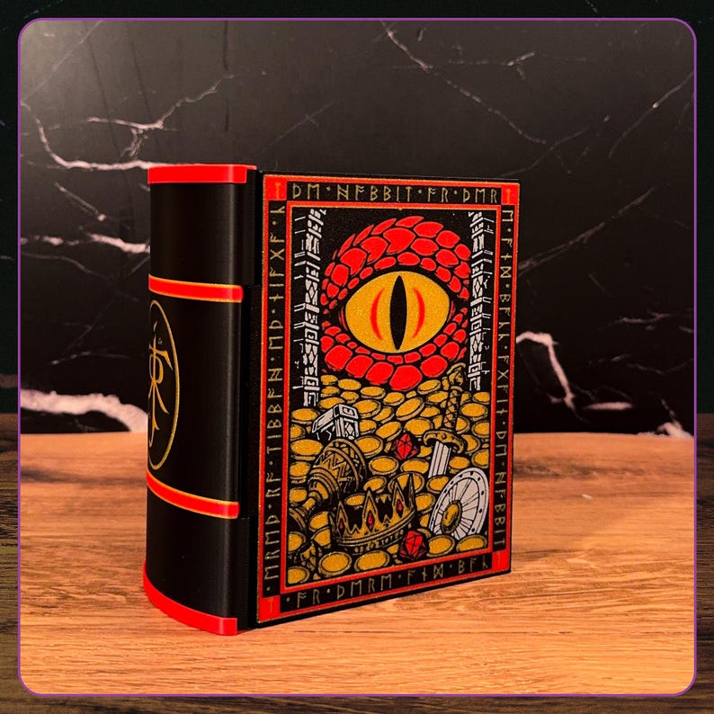 May include: A black rectangular box shaped like a book with red accents. The front panel features a large red dragon eye surrounded by gold coins, a crown, a sword, and a shield. The border contains text reading DM N FBBIT FR DMR.