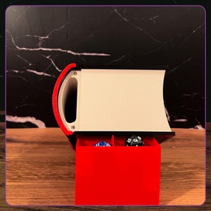 May include: A black and white book-shaped storage container with a red pull-out drawer featuring two compartments. Inside the drawer are two multi-sided gaming dice, one blue and one black, resting within the red plastic structure.