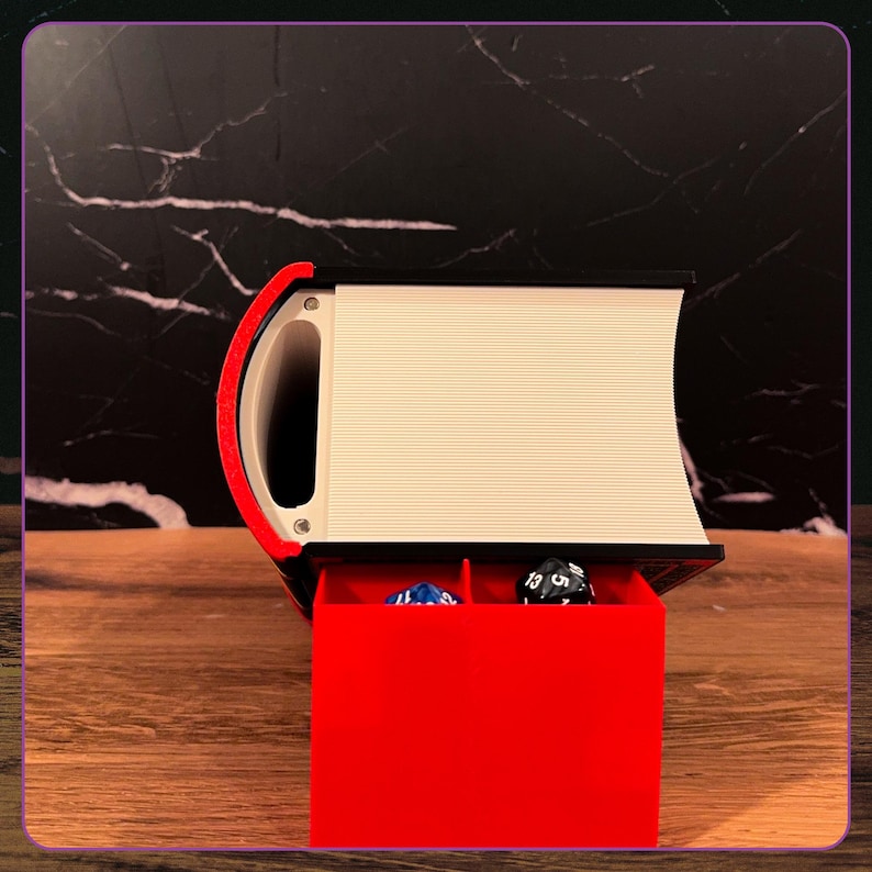 May include: A black and white book-shaped storage container with a red pull-out drawer featuring two compartments. Inside the drawer are two multi-sided gaming dice, one blue and one black, resting within the red plastic structure.