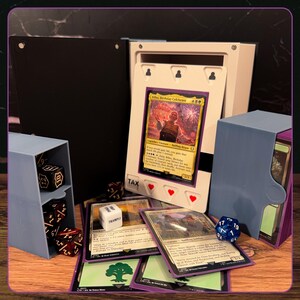 May include: A black and white card display frame holding a Bilbo, Birthday Celebrant trading card, accompanied by a light blue dice storage tower, a light blue deck box, several black and red dice, a white six-sided die, and multiple trading cards.