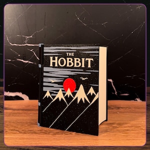 May include: A hardcover book titled The Hobbit with a black cover featuring a graphic illustration of white mountain peaks, a bright red sun, and two small birds flying against a dark sky. The book has a black spine and white pages.