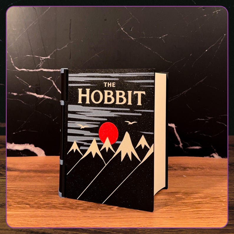 May include: A hardcover book titled The Hobbit with a black cover featuring a graphic illustration of white mountain peaks, a bright red sun, and two small birds flying against a dark sky. The book has a black spine and white pages.