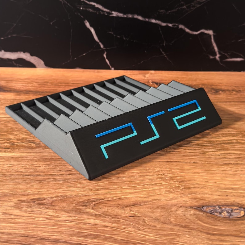 May include: A black and grey PS2 game controller stand featuring a blue and teal PS2 logo. The stand has a tiered design with slots for controllers and accessories. The stand is made of plastic and sits on a wooden surface.
