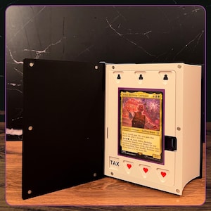 May include: A white and black display case for a trading card, featuring a hinged door with magnetic closures. The interior holds a Bilbo, Birthday Celebrant card. The white frame includes three red heart icons and the text TAX printed in black.