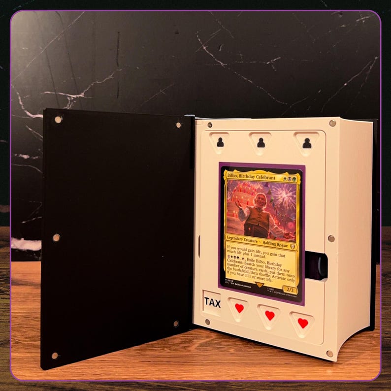 May include: A white and black display case for a trading card, featuring a hinged door with magnetic closures. The interior holds a Bilbo, Birthday Celebrant card. The white frame includes three red heart icons and the text TAX printed in black.