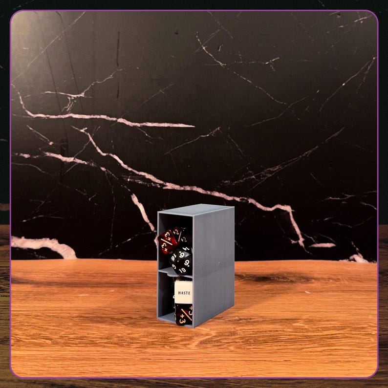 May include: A grey, rectangular, two-tiered storage organizer made of plastic. The top shelf contains several multi-sided gaming dice in red and black. The bottom shelf holds a white rectangular token with the word HASTE printed in black.
