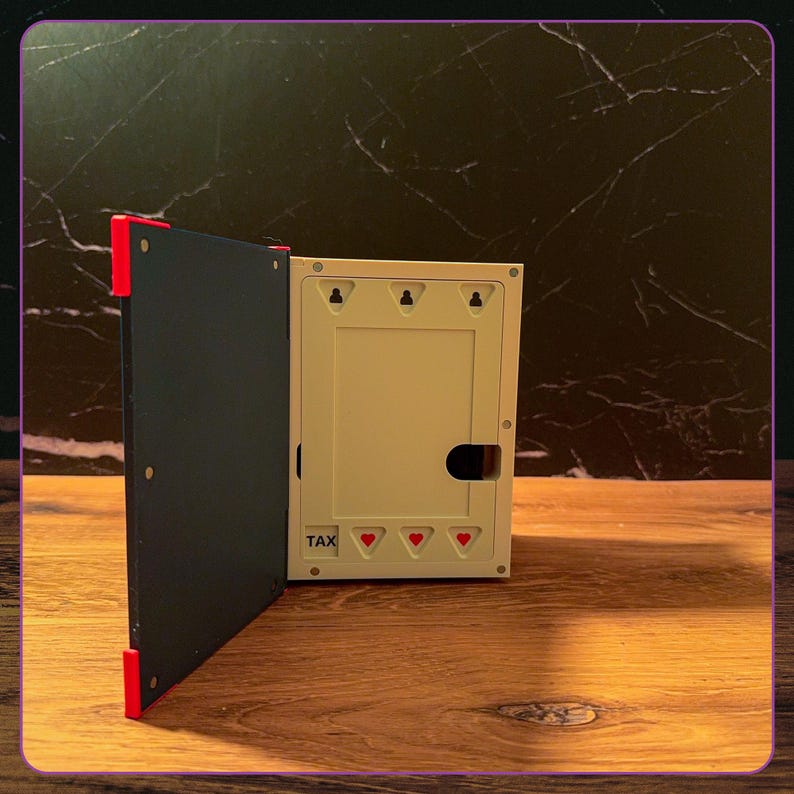 May include: A rectangular box with a dark blue hinged cover featuring red corner accents. The interior is off-white with three red heart icons and the word TAX printed in black. The structure includes three keyhole slots for wall mounting.