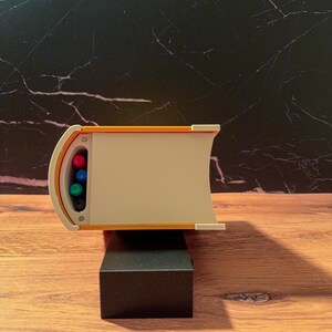 May include: A horizontal, book-shaped desk organizer with a cream-colored spine and orange top and bottom edges. The left side features a recessed, curved opening containing four colored markers in pink, blue, green, and black, arranged vertically.
