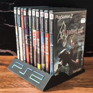 May include: A black PlayStation 2 game holder with a blue and teal PS2 logo, holding several PlayStation 2 game cases. The game cases include titles like "Resident Evil 4", "Guitar Hero", and "Final Fantasy X".