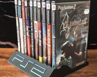 May include: A black PlayStation 2 game holder with a blue and teal PS2 logo, holding several PlayStation 2 game cases. The game cases include titles like "Resident Evil 4", "Guitar Hero", and "Final Fantasy X".