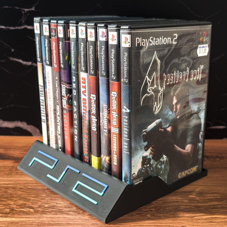 May include: A black PlayStation 2 game holder with a blue and teal PS2 logo, holding several PlayStation 2 game cases. The game cases include titles like "Resident Evil 4", "Guitar Hero", and "Final Fantasy X".