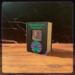 May include: A black rectangular deck box with a light-colored spine, featuring the green text Witherbloom Pestilence above a square cutout displaying a trading card. A purple and green circular emblem is printed below the cutout, with purple corner accents.
