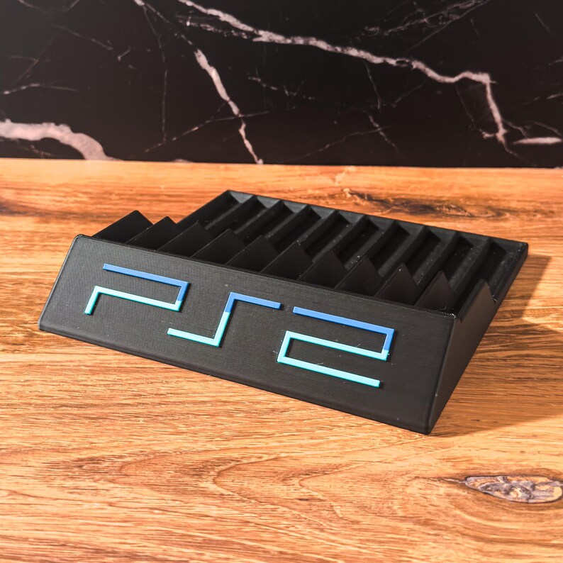 May include: Black PS2 game holder with a blue and light blue "PS2" logo. The holder has multiple slots for game storage and is made of a solid material. The holder is on a wooden surface.
