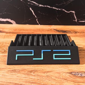 May include: A black PS2 game console stand with blue detailing. The stand has vertical slots for game storage and features the PS2 logo in blue. The stand is made of a matte black material and sits on a wooden surface.