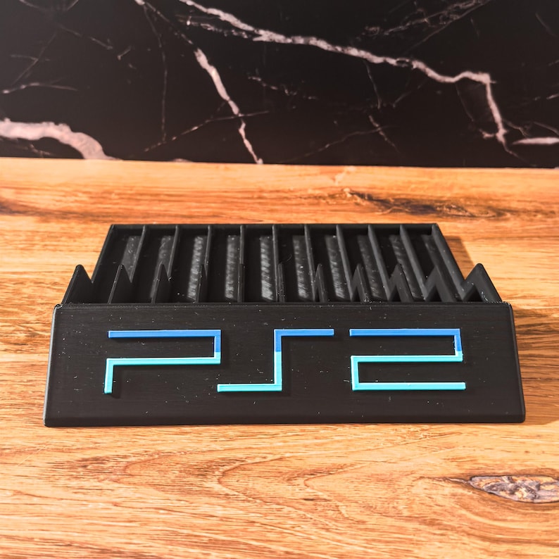 May include: A black PS2 game console stand with blue detailing. The stand has vertical slots for game storage and features the PS2 logo in blue. The stand is made of a matte black material and sits on a wooden surface.