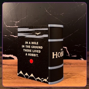 May include: A black book cover featuring the text 'IN A HOLE IN THE GROUND THERE LIVED A HOBBIT.' with a white bird, a red circle, and white mountain silhouettes at the bottom. The spine is black with grey horizontal bands and the text 'THE HOBBIT'.