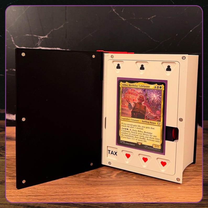 May include: A white and black display case for a trading card, featuring a magnetic closure and a central slot holding a Bilbo, Birthday Celebrant Magic: The Gathering card. The case includes the text TAX and three red heart-shaped cutouts.