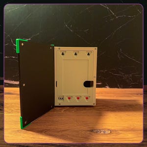 May include: A white rectangular wall-mounted box with a black hinged door featuring bright green corner accents. The interior panel displays the word TAX in black text above three small red heart icons, with three keyhole slots at the top.