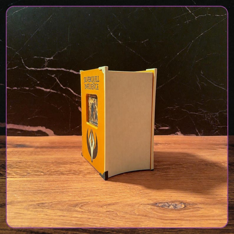 May include: A rectangular wooden deck box with a yellow front panel featuring the text Silverquill Influence and a black and white emblem. The box has a cutout window displaying a trading card, light wood side panels, and small black corner accents.