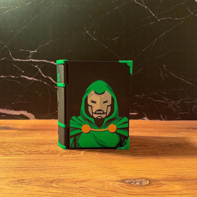 May include: A rectangular black storage box featuring a graphic illustration of a character wearing a green hooded cloak and a metallic mask. The box has bright green corner accents and a black spine with green detailing, presenting a comic book aesthetic.