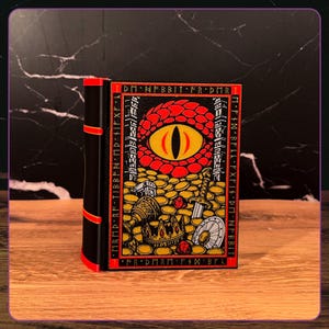 May include: A black and red book-shaped box featuring a graphic illustration of a large dragon eye overlooking a pile of gold coins, a crown, a sword, and a shield. The border contains runic-style text, and the spine has three red horizontal bands.