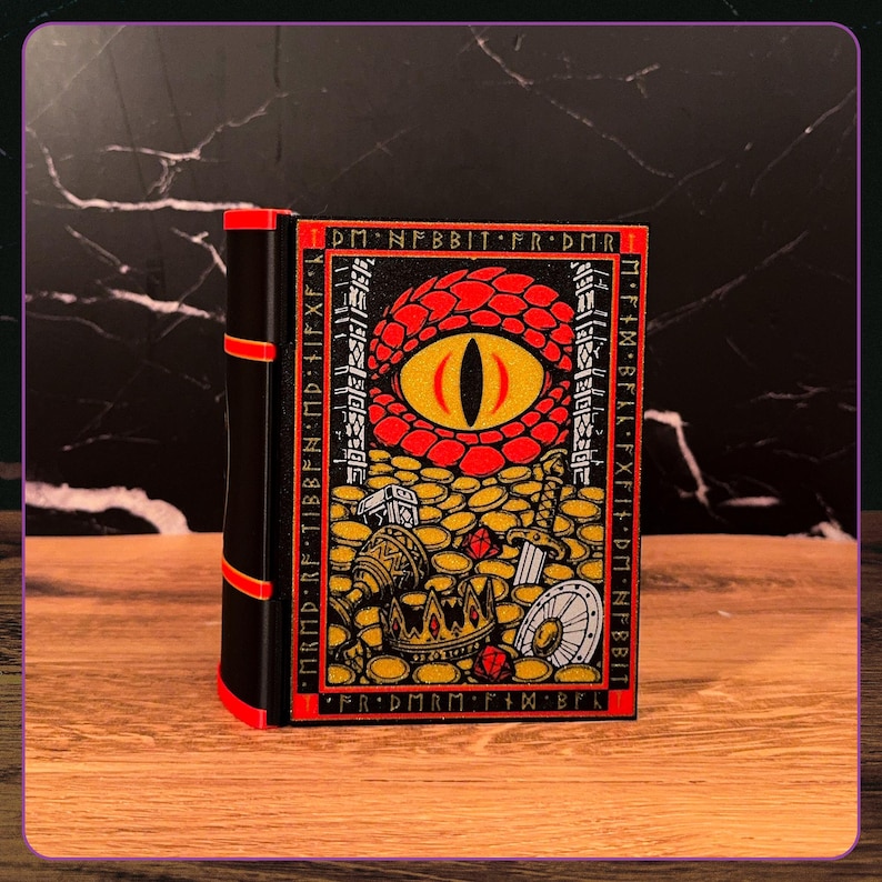 May include: A black and red book-shaped box featuring a graphic illustration of a large dragon eye overlooking a pile of gold coins, a crown, a sword, and a shield. The border contains runic-style text, and the spine has three red horizontal bands.
