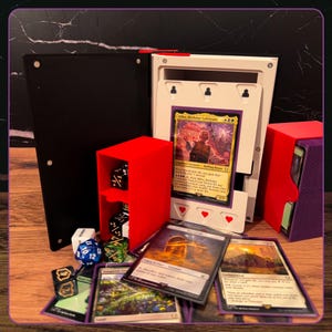 May include: A black and white card storage box with a red dice holder containing black dice, a purple deck box, and several trading cards including a Bilbo, Birthday Celebrant card. The setup includes various polyhedral dice scattered around.