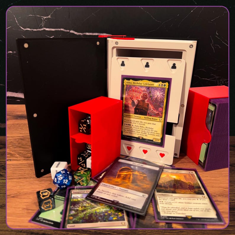 May include: A black and white card storage box with a red dice holder containing black dice, a purple deck box, and several trading cards including a Bilbo, Birthday Celebrant card. The setup includes various polyhedral dice scattered around.