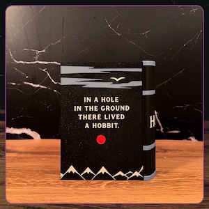 May include: A black rectangular book cover featuring the white text IN A HOLE IN THE GROUND THERE LIVED A HOBBIT. The design includes a small white bird, a red circular sun, and white mountain silhouettes at the bottom with a light blue spine.