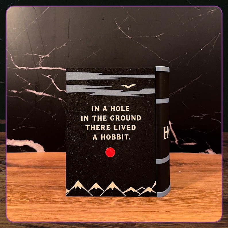 May include: A black rectangular book cover featuring the white text IN A HOLE IN THE GROUND THERE LIVED A HOBBIT. The design includes a small white bird, a red circular sun, and white mountain silhouettes at the bottom with a light blue spine.