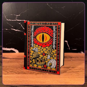 May include: A black book with a red spine and a front cover illustration featuring a large red dragon eye surrounded by gold coins, a crown, a sword, and a shield. The border contains the text DM NFBBIT FR DMR and other runic characters.