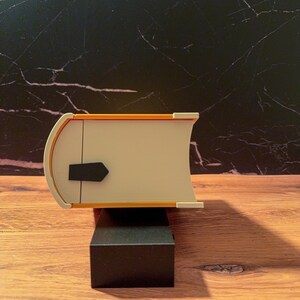 May include: A rectangular object shaped like a closed book with a cream-colored spine and cover, featuring a bright orange border and a small black tab on the side. The object is positioned horizontally on a black rectangular base.
